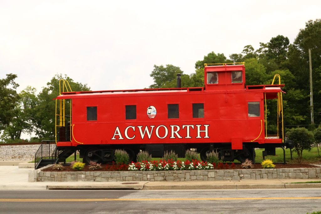 Bright red train car with Acworth in white letters painted on the side parked in a flower bed.