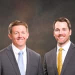 Two men wearing suits standing in front of a professional photographers backdrop.