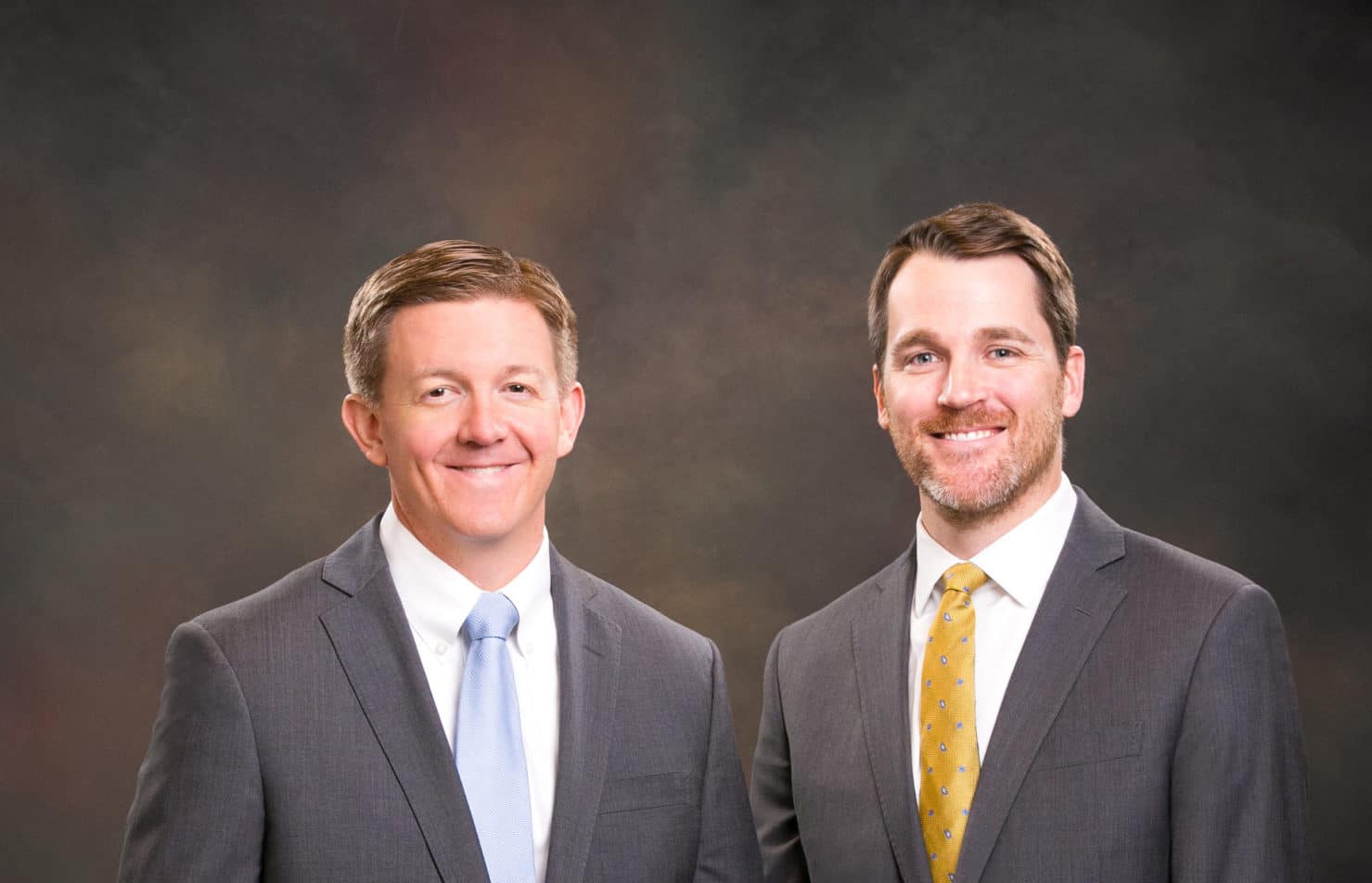 Two men wearing suits standing in front of a professional photographers backdrop.