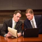 Professional male attorneys at a desk, conversing about something on a legal notepad, with a laptop in front of them.