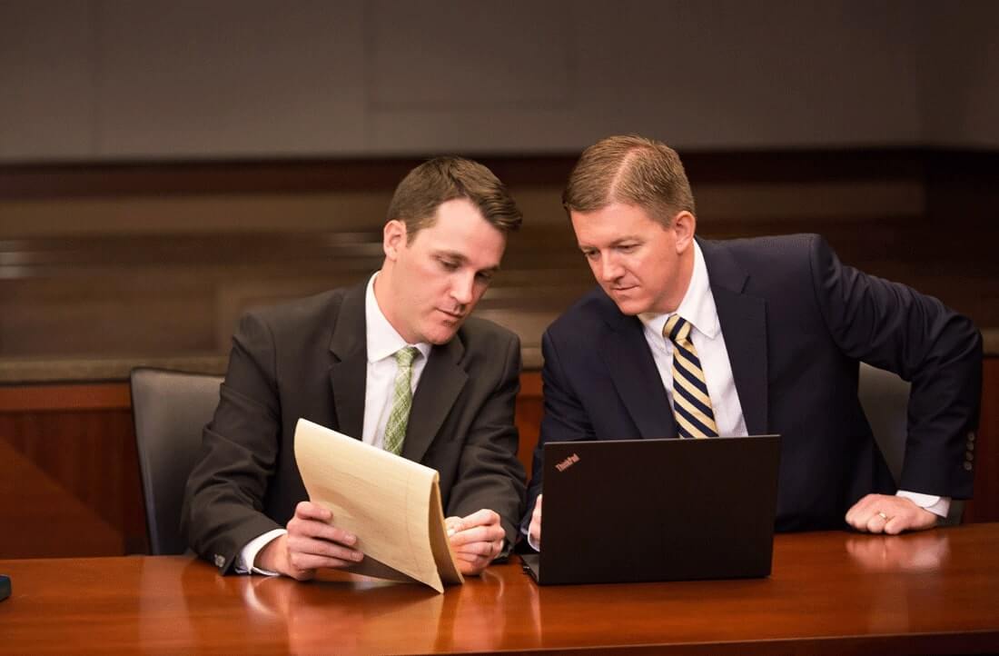 Professional male attorneys at a desk, conversing about something on a legal notepad, with a laptop in front of them.