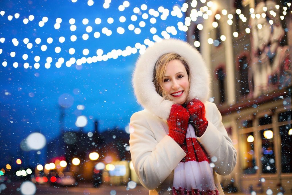 A woman wearing a white coat with a fur lined hood wrapped around her face with bright red gloves on, standing outside with snow falling around her and strings of lights blurred in the backgound.