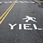 A stick figure painted on a road, walking across it about the word YIELD.