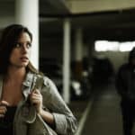 A scared girl walking alone in a parking deck looking over her shoulder at the man in the wearing a black hooded jacket walking behind her.