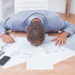 A man laying his head down at a work desk with papers, a laptop, and other work tools surrounding him.