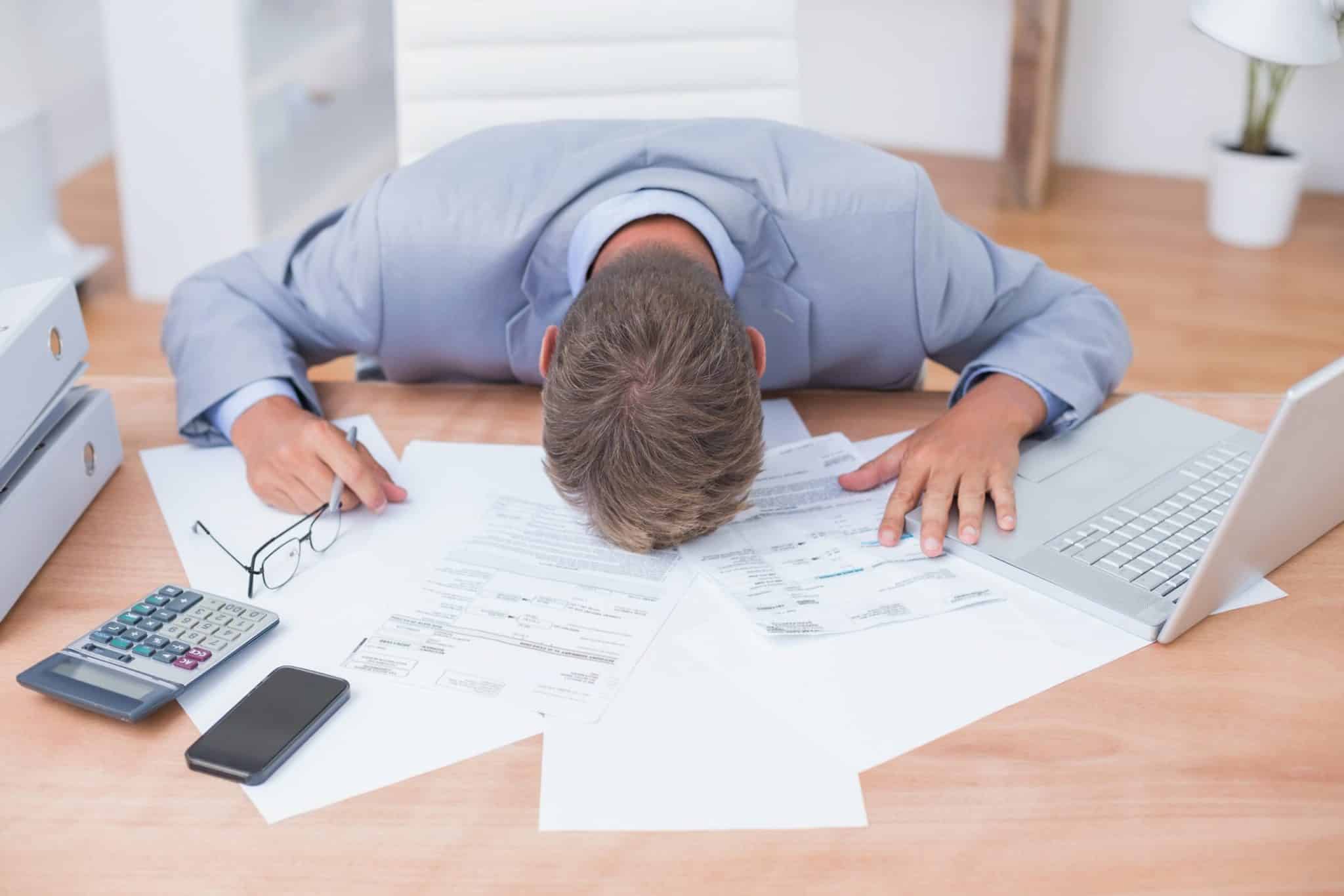 A man laying his head down at a work desk with papers, a laptop, and other work tools surrounding him.