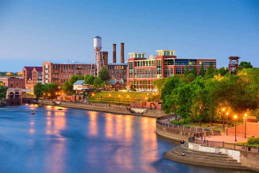 Large brick buildings beside a river lined by a concrete retaining wall and sidewalk.