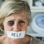 An elderly woman with a black eye and white tape covering her mouth with the word help written on the tape.