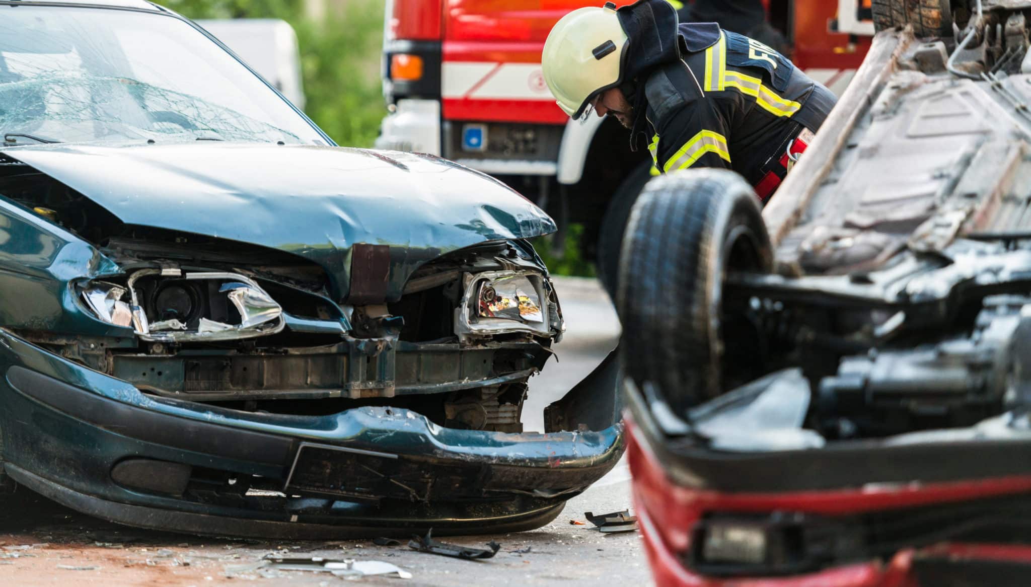 Multiple car wreck, the front end of a dark green car smashed and a red car flipped upside down with a firemen assesing the damage.