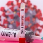 A large syringe and a citrate tube both labeled COVID-19 in front of a blurry view of a COVID-19 molecule.