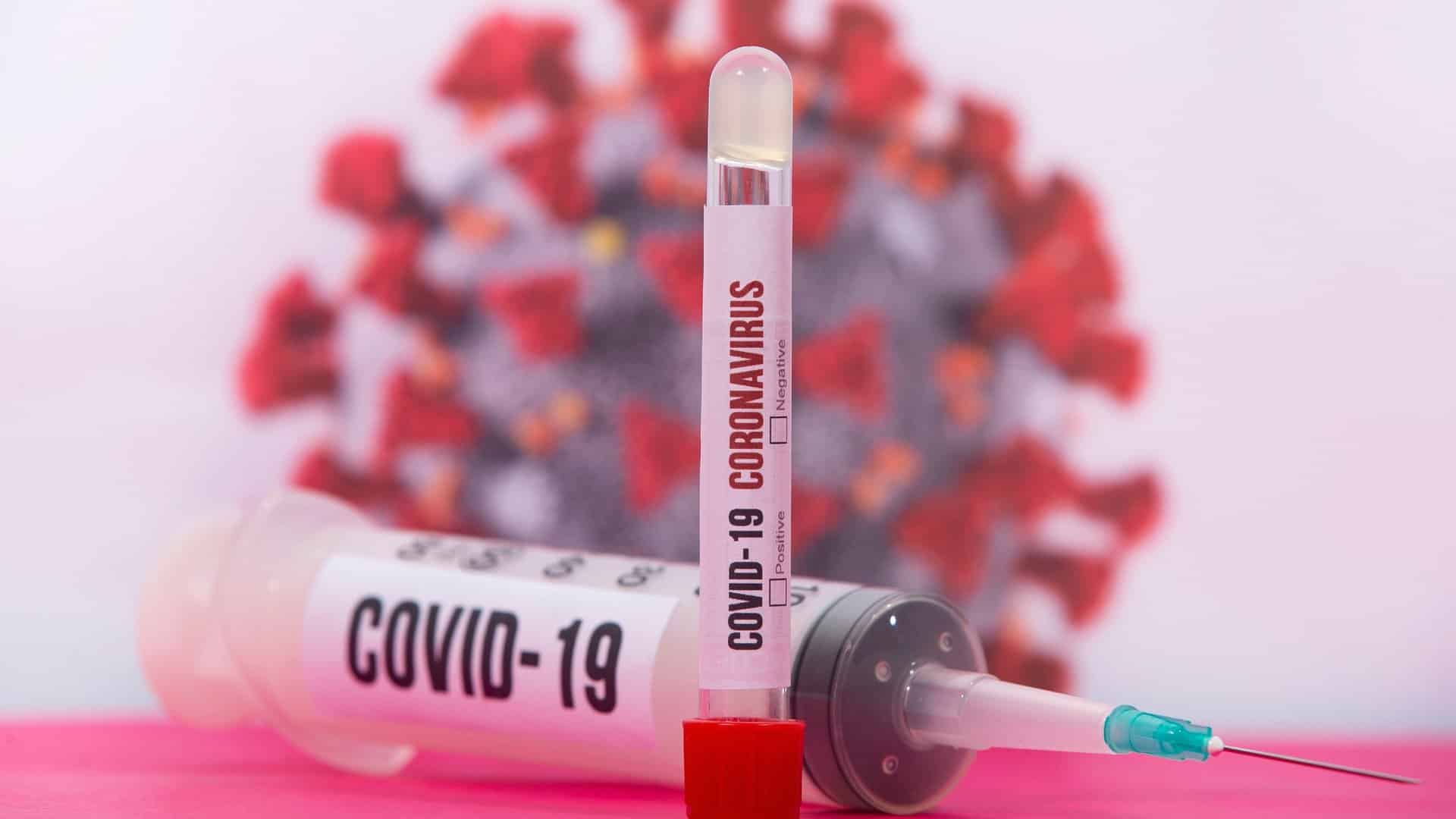 A large syringe and a citrate tube both labeled COVID-19 in front of a blurry view of a COVID-19 molecule.