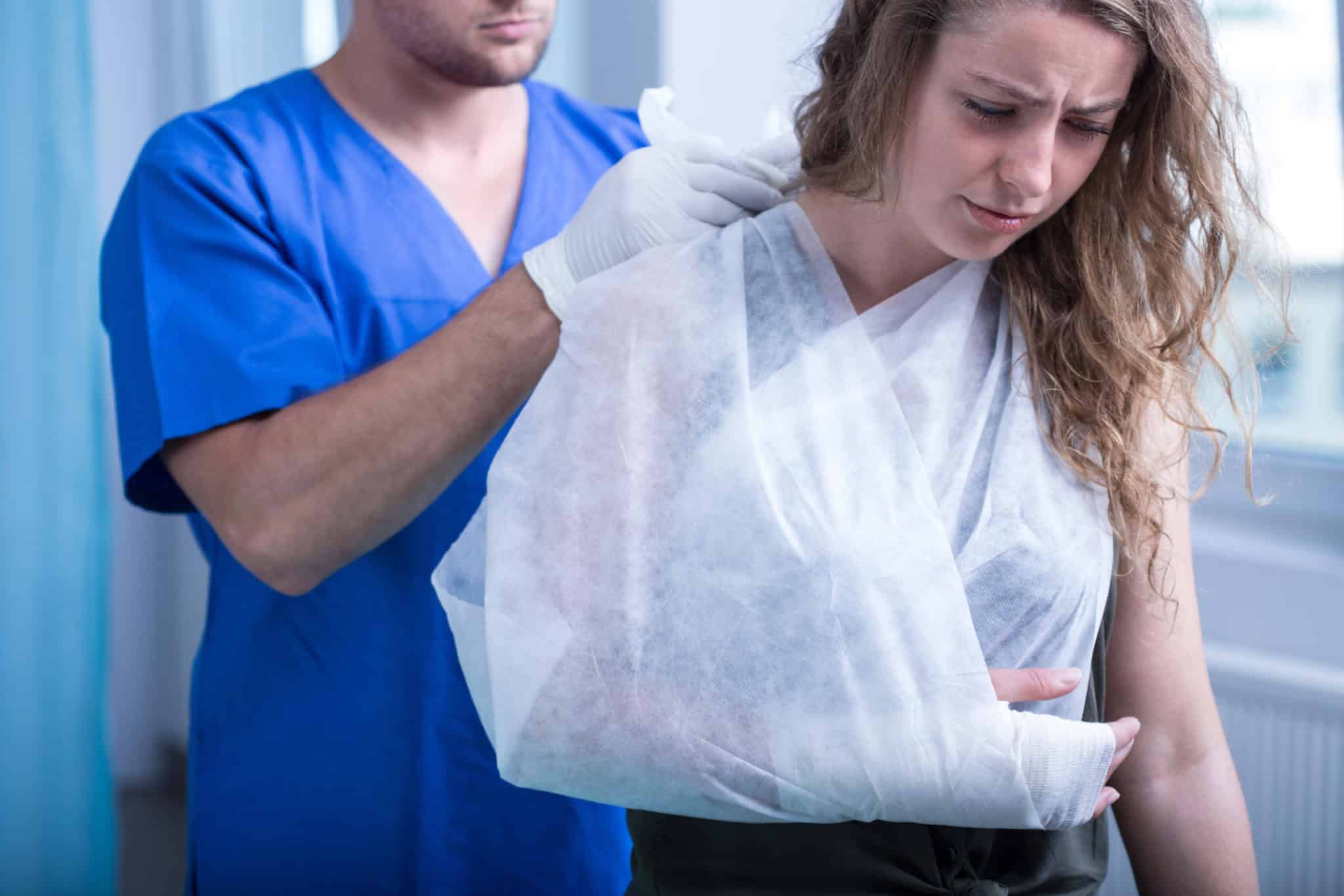 A doctor wearing blue scrubs tieing a white sling around a woman's right arm.