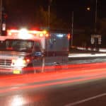 An ambulance with it's lights on parked on the side of the road at night with the blurred lights of cars driving by.