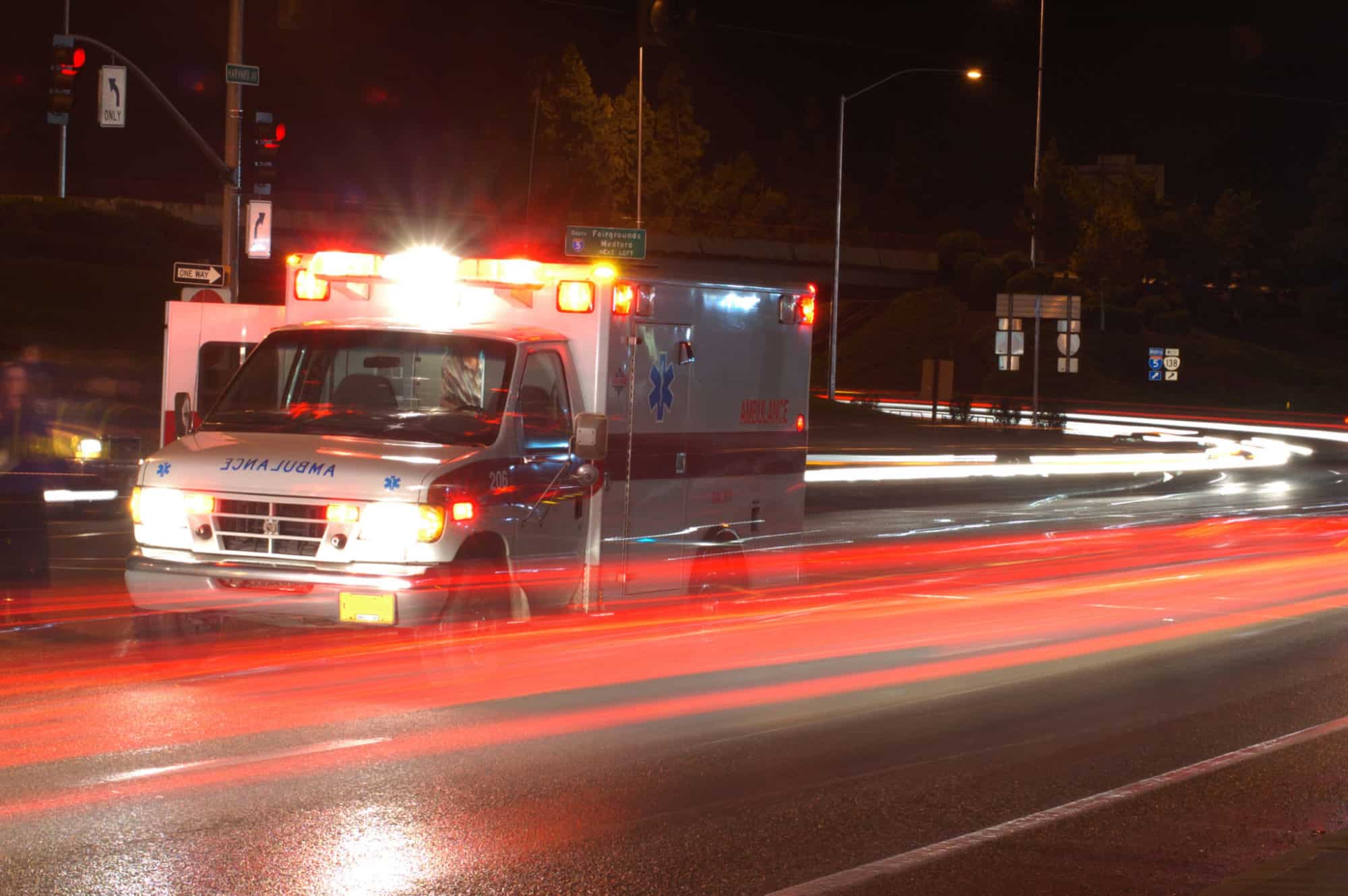 An ambulance with it's lights on parked on the side of the road at night with the blurred lights of cars driving by.