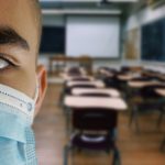 Close-up of a kids face wearing a blue face mask in front of a blurry view of a classroom in the background.