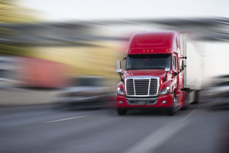 A bright red semi truck pulling a white trailer surrounded by a blurry background.