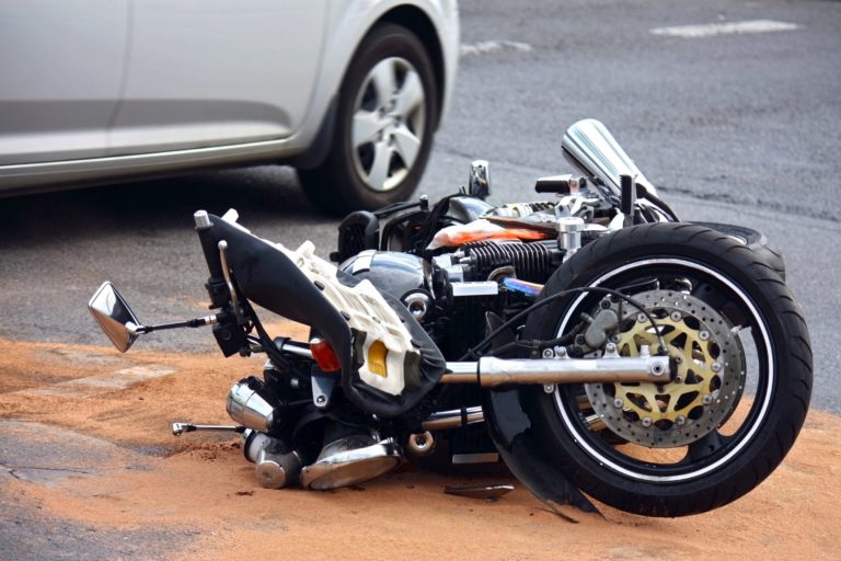 Close-up of a wrecked motorcycle on a road with a silver car in the background.