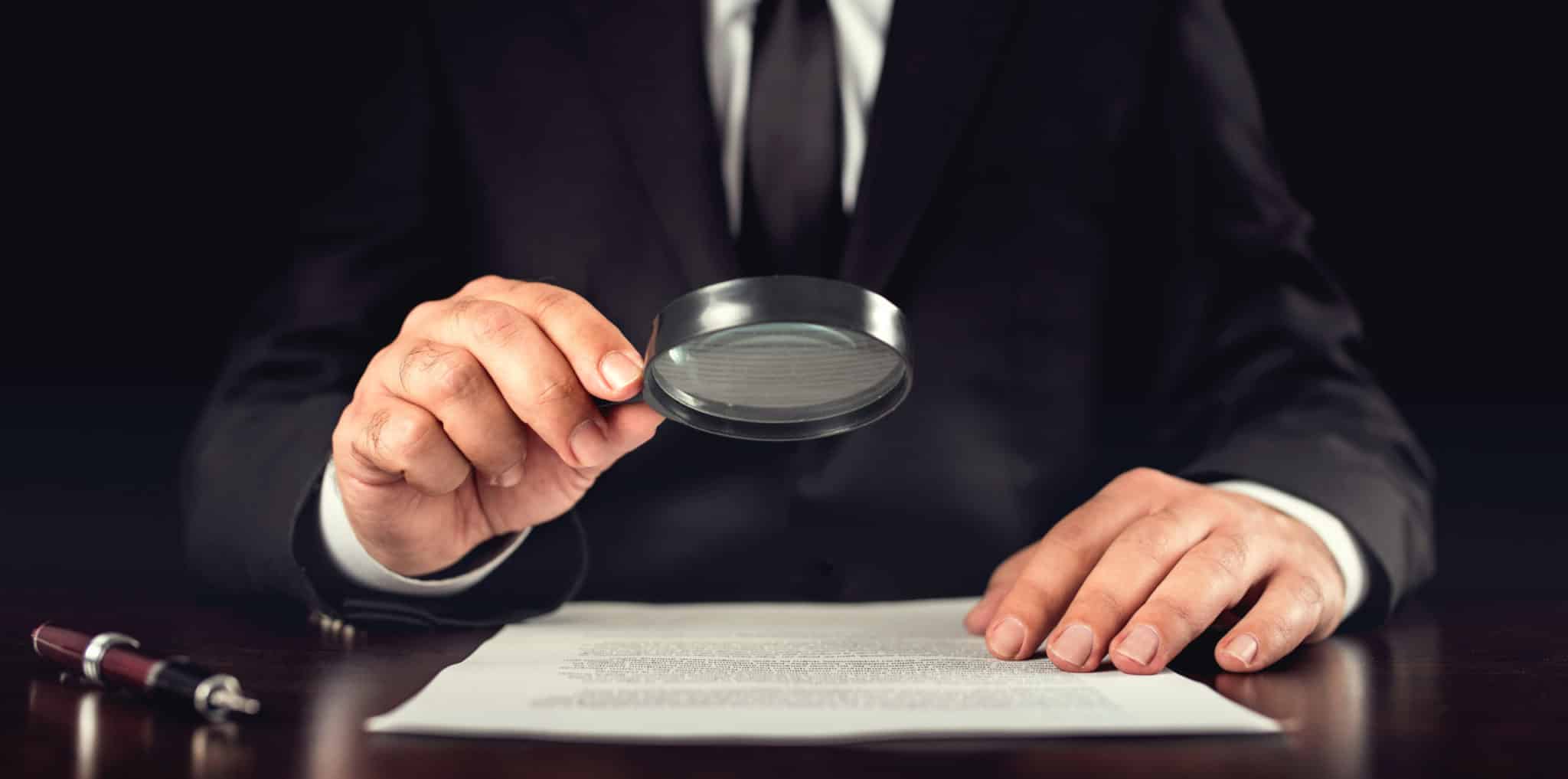 Man in professional suit holding a magnifying glass over a document.