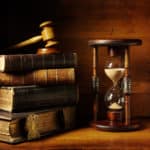 A stack of four old hardcover books with a wooden gavel on top, beside a large hour glass.