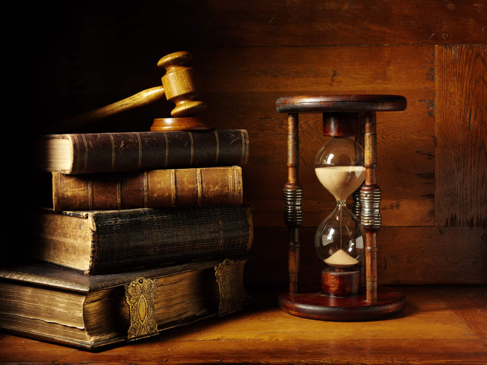 A stack of four old hardcover books with a wooden gavel on top, beside a large hour glass.