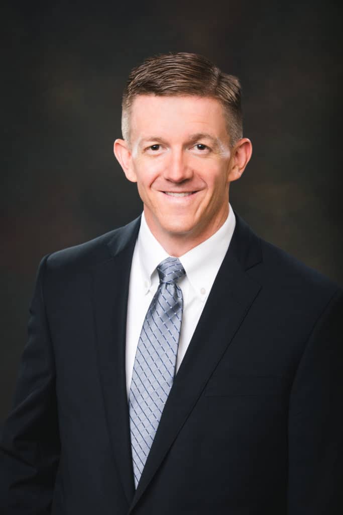 A headshot of a male professional wearing a navy blue suit, white shirt and shiny blue tie.