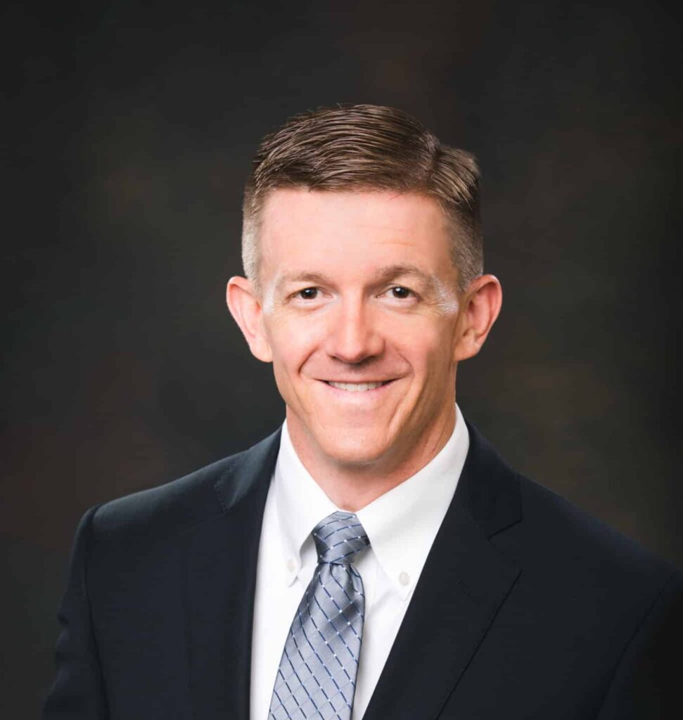 A headshot of a male professional wearing a navy blue suit, white shirt and shiny blue tie.