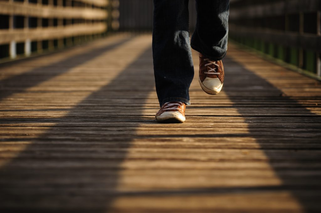 A person wearing blue jeans and brown tennis-shoes walking toward the camera, across a bridge.