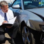 A man wearing a suit, crouched down, beside a silver car with frontend damage, writing in a notebook.