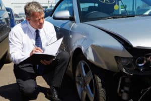 A man wearing a suit, crouched down, beside a silver car with frontend damage, writing in a notebook.