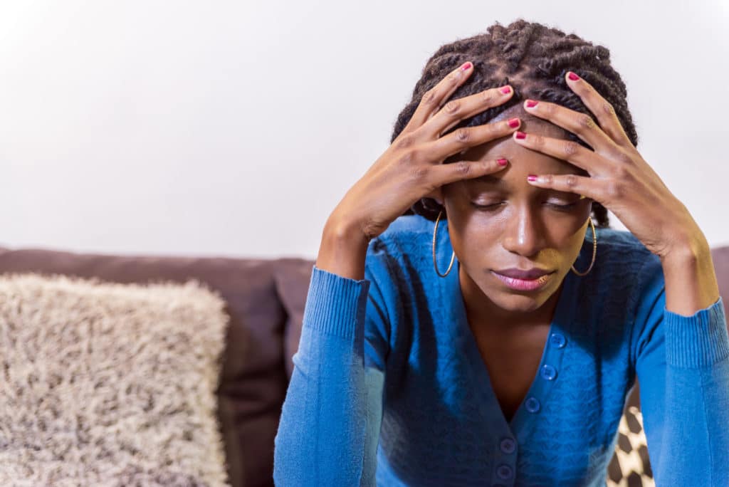 An African American woman sitting on a couch with her head in her hands appearing stressed.