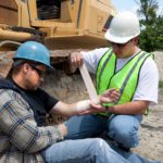 One construction worker wrapping an injured arm of another construction worker with construction equipment in the background.