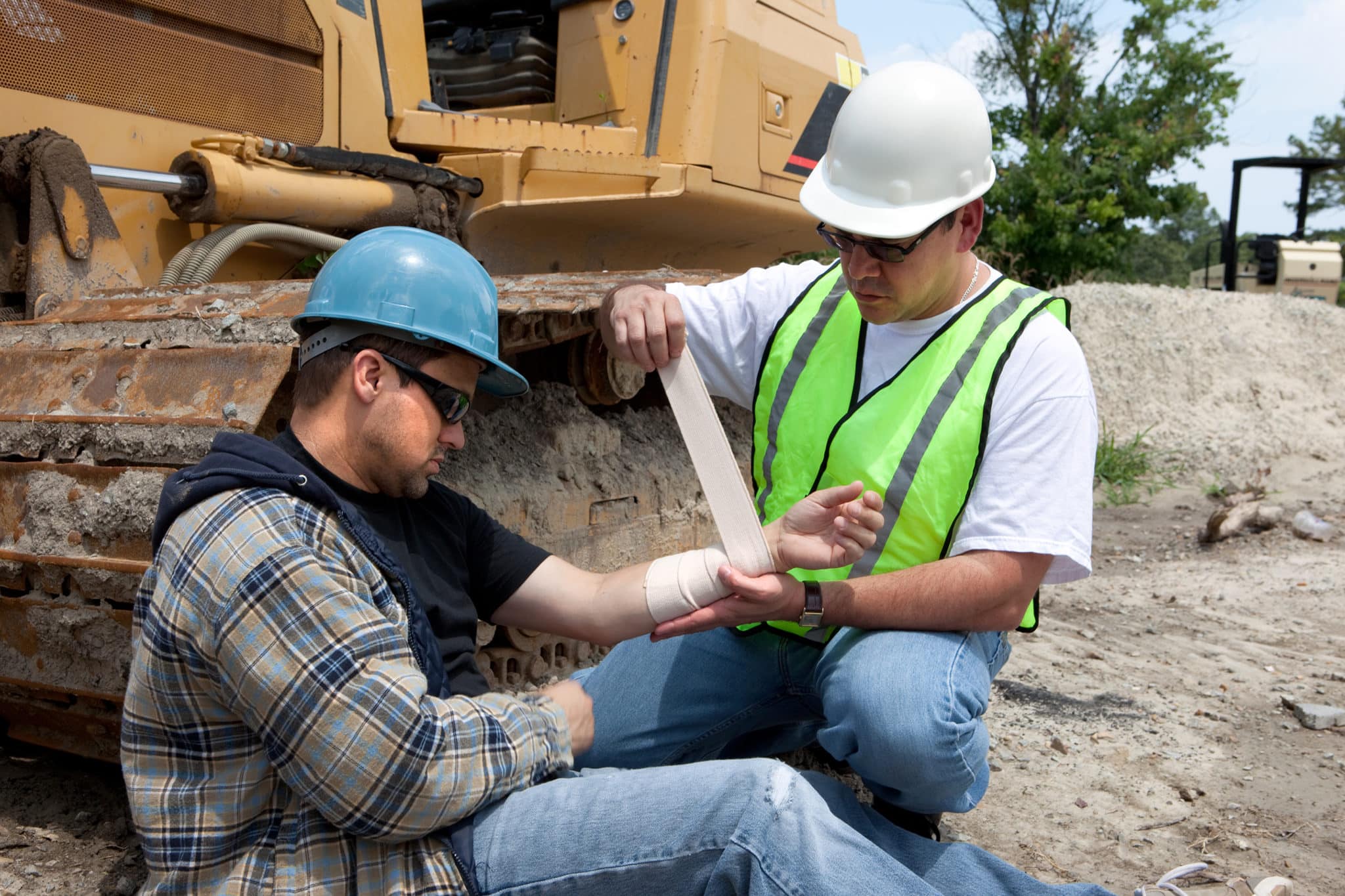 One construction worker wrapping an injured arm of another construction worker with construction equipment in the background.