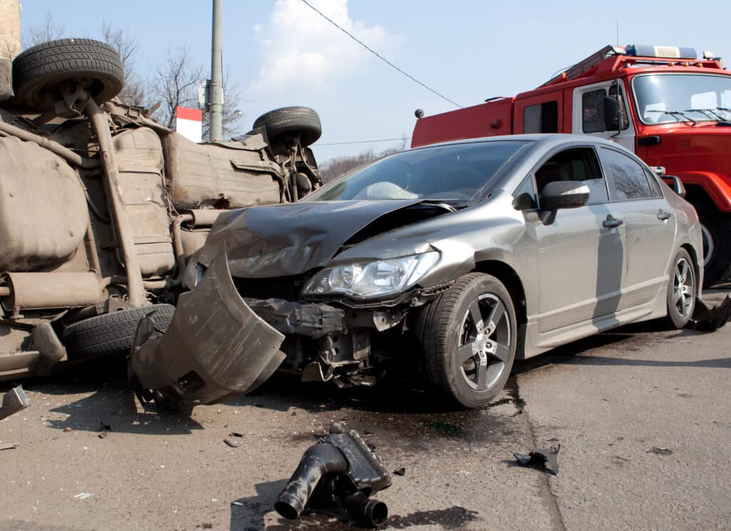 The front end of a car is smashed after a car wreck. The underside of another car lying on it's side is visible in the background.