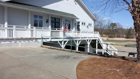 There is a railing beside a paved concrete slab in front of a white building with a deck.