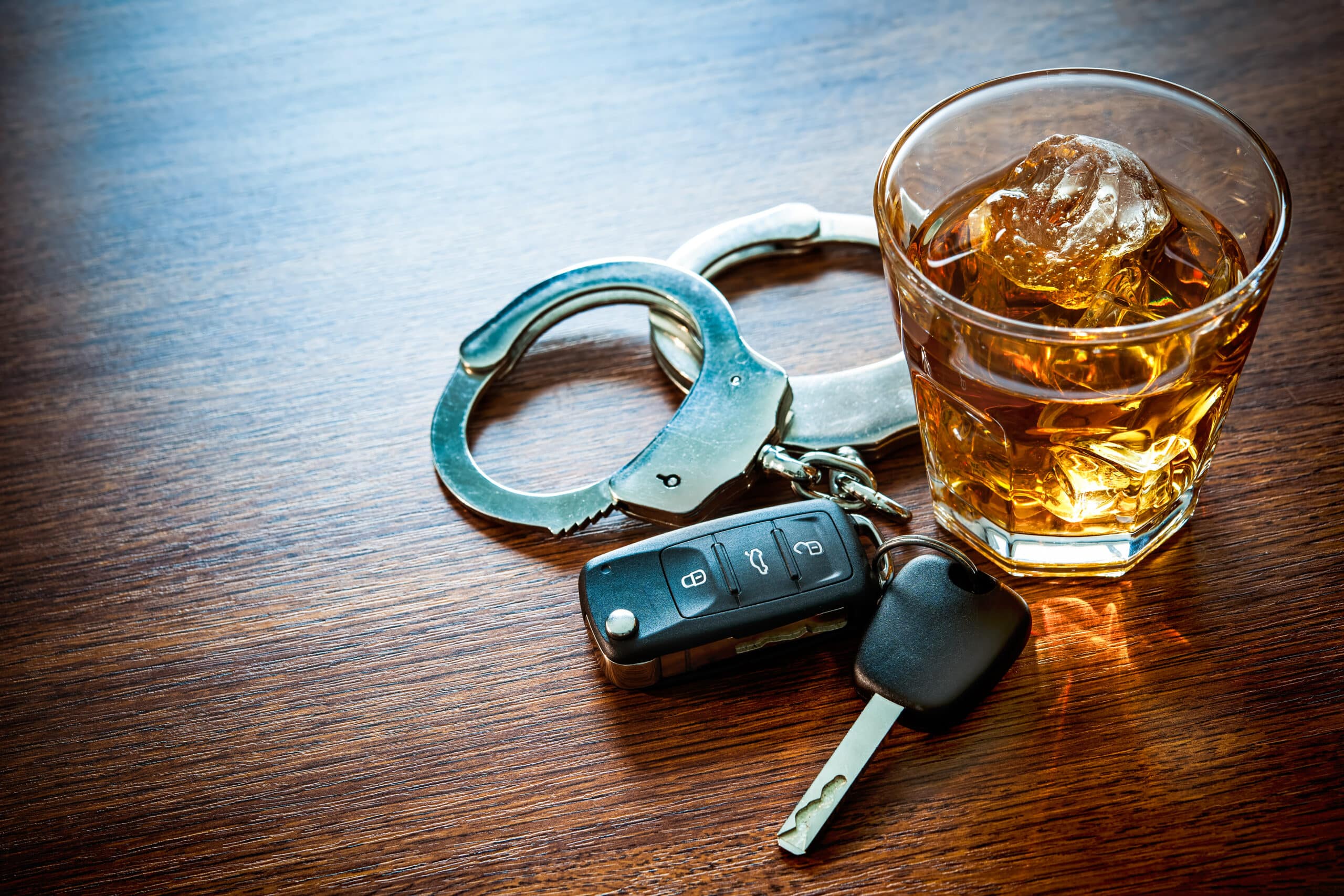 A clear glass of liquor on ice beside a set of car keys and handcuffs on a wooden table.