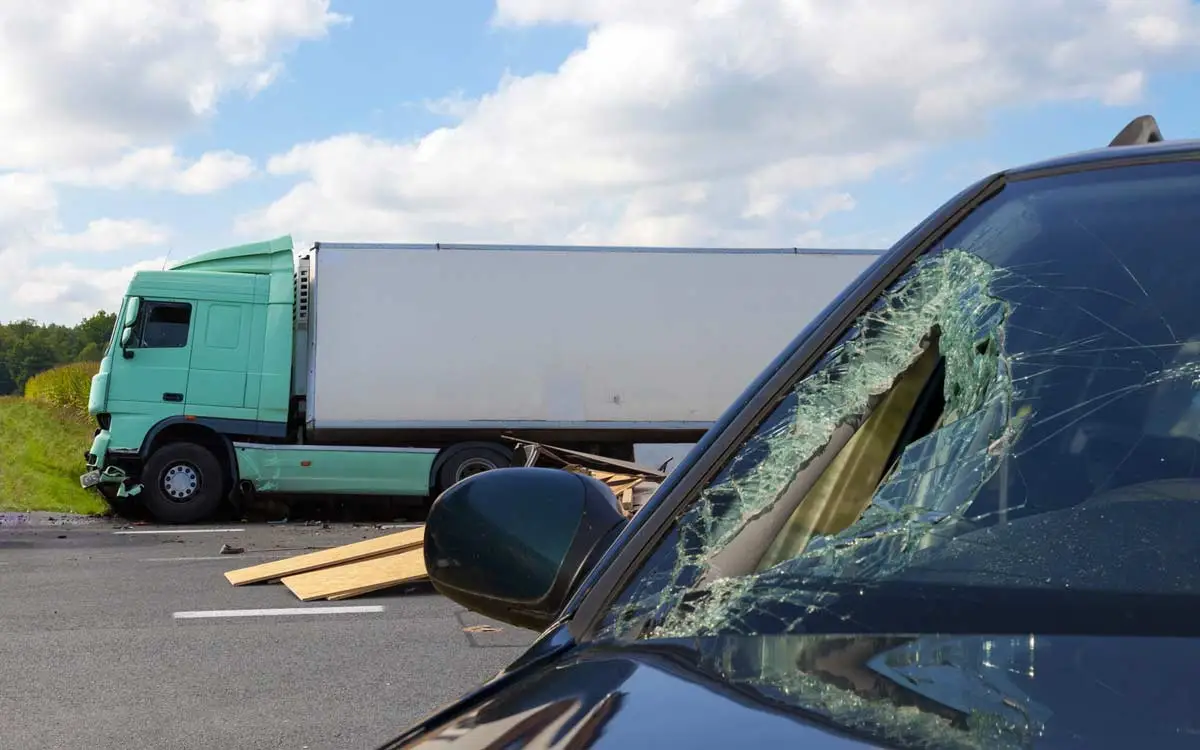 Car wreck scene with a tractor trailer truck carrying lumber and a small car with cracked windshield.