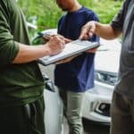 Three Asian men meet with an insurance adjuster to discuss car insurance policies, reviewing premiums, deductibles, coverage, and a recent claim settlement.