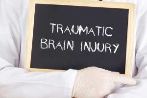 A close-up of someone's chest wearing a white coat and white surgical gloves with a stethoscope around their neck, holding a black chalkboard with the words TRAUMATIC BRAIN INJURY written on it.